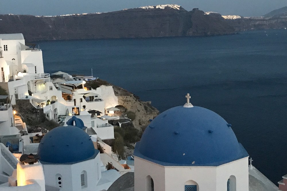 The blue domes of Oia at dusk, with the caldera and the lit cliffs of Santorini behind, photographed by Michael Olaiya