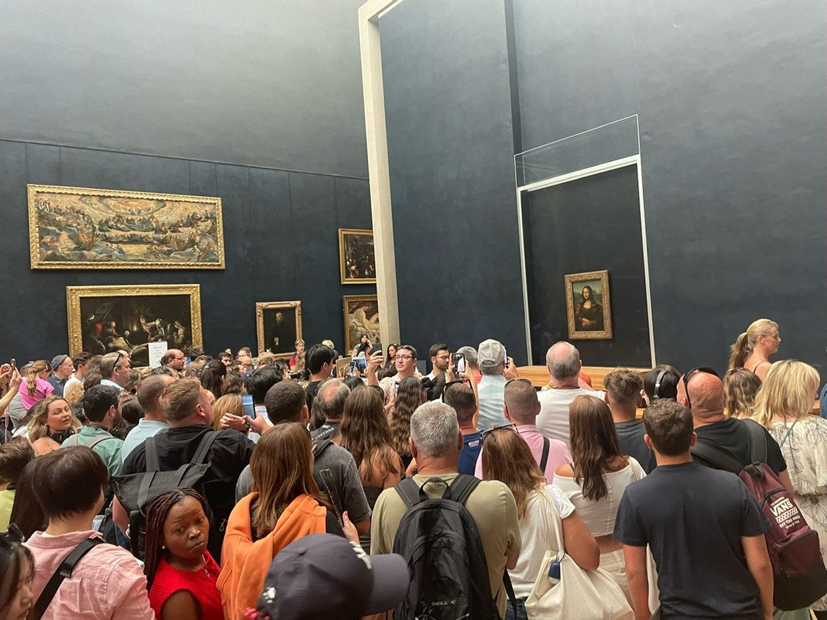 The Mona Lisa, small on a far wall, above a packed room of visitors holding up phones